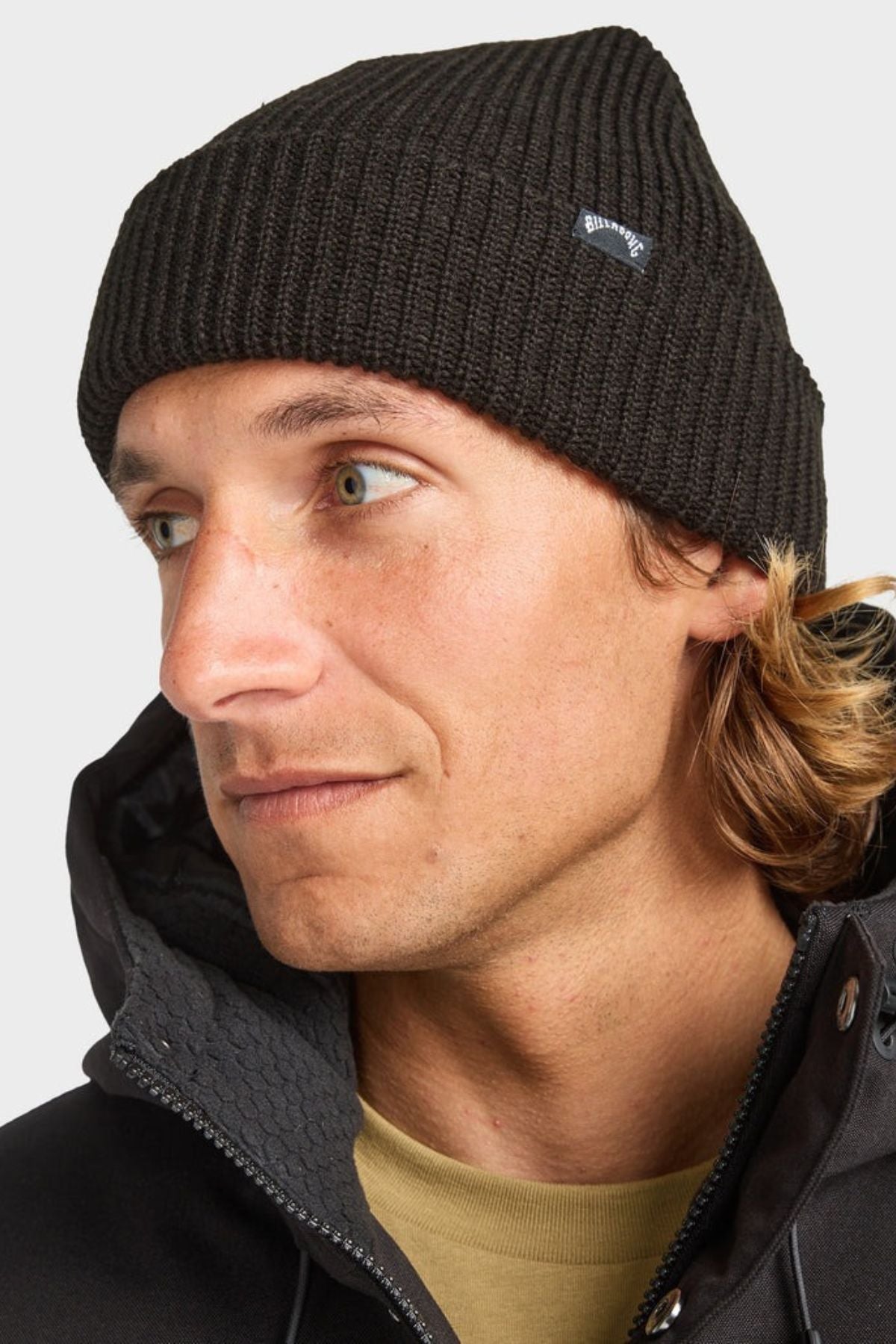 A man with light skin and green eyes wears a black Billabong Arcade Beanie and a black jacket, looking to the side against a plain light background.