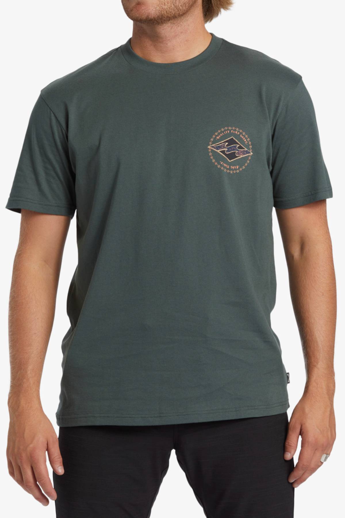 A person with light skin and shoulder-length light brown hair is wearing a dark green Billabong Rotar Diamond T-Shirt, which has a small geometric logo pattern on the left chest area. The regular fit tee is made of 100% cotton. They are also dressed in black pants or shorts.
