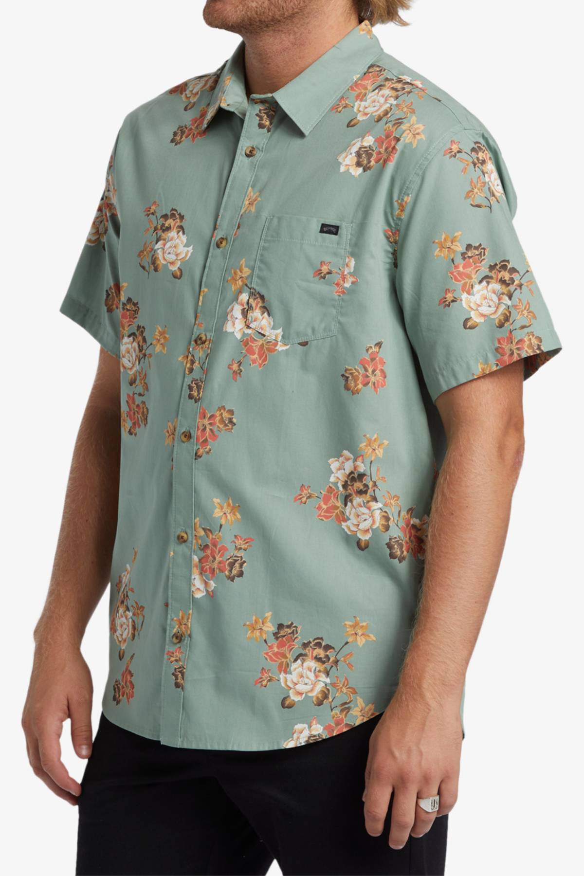A person stands sideways with one hand in their pocket, wearing a light green Billabong Sundays Mini Short Sleeve Shirt featuring a bold orange, yellow, and white seasonal print.