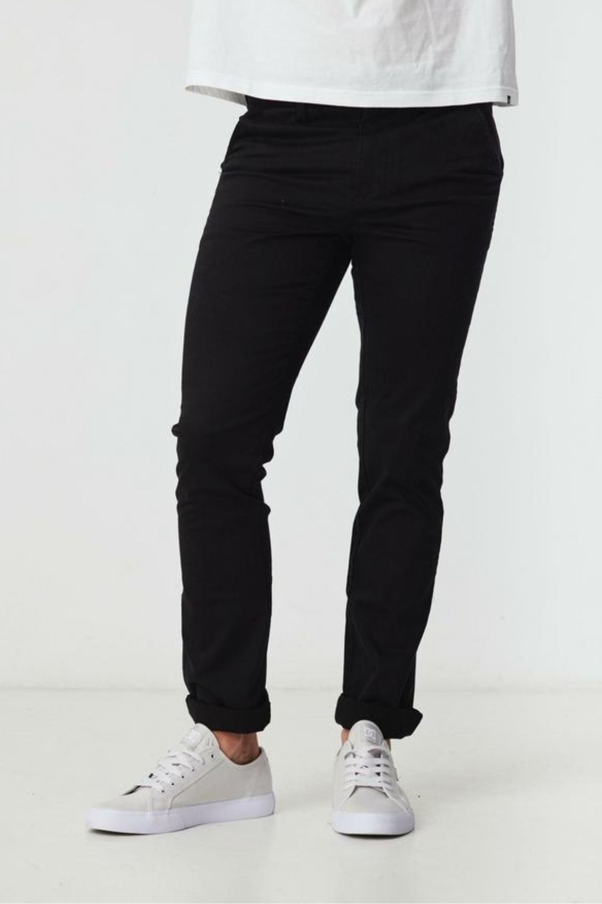 A person stands against a plain white background wearing a white t-shirt, BLKWD Clay Slim Chino Pant in black with cotton stretch fabric, and white sneakers. Their upper body is partially out of the frame.