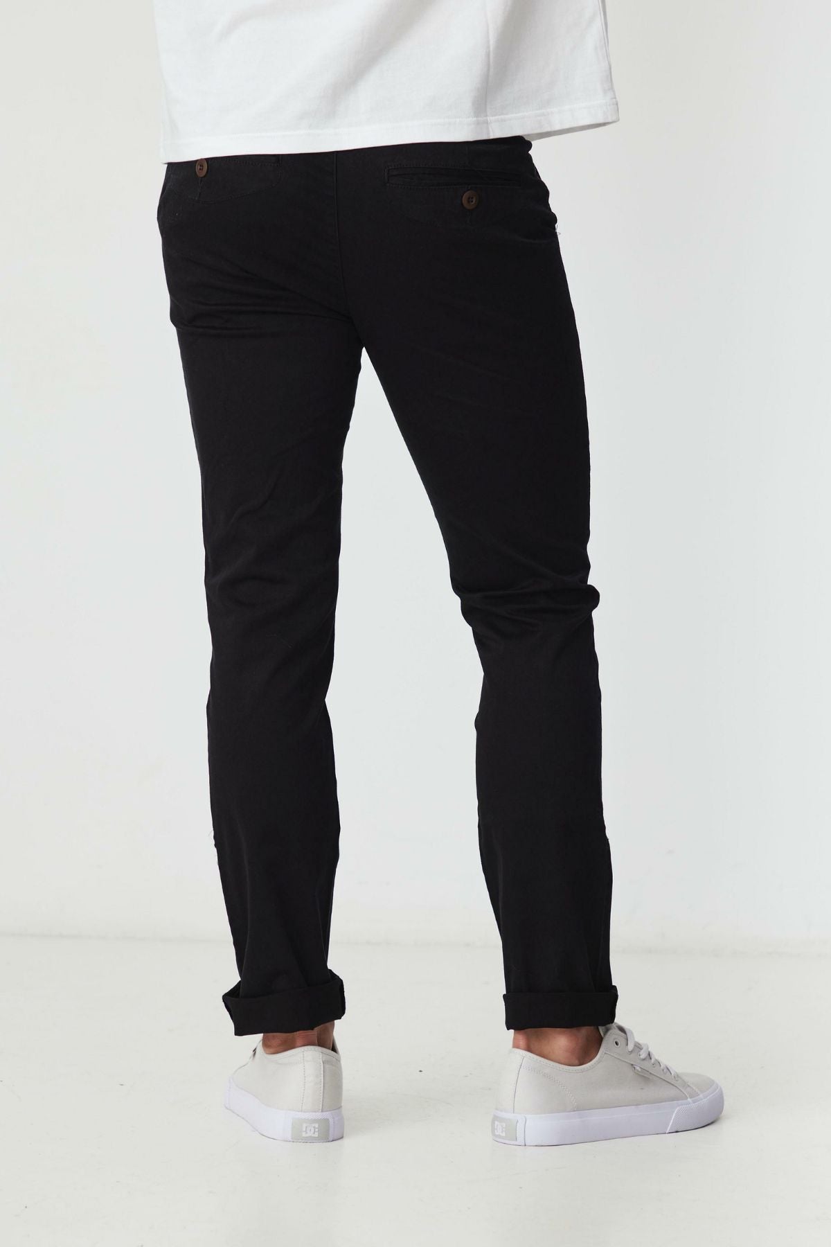 A person stands facing away, wearing BLKWD Clay Slim Chino Pant in black, made from cotton stretch fabric, paired with a white shirt and white sneakers against a plain white background.