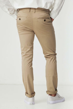 A person wearing BLKWD Clay Slim Chino Pant, made from cotton stretch fabric in light brown, stands with hands in back pockets and faces away from the camera against a plain white background.