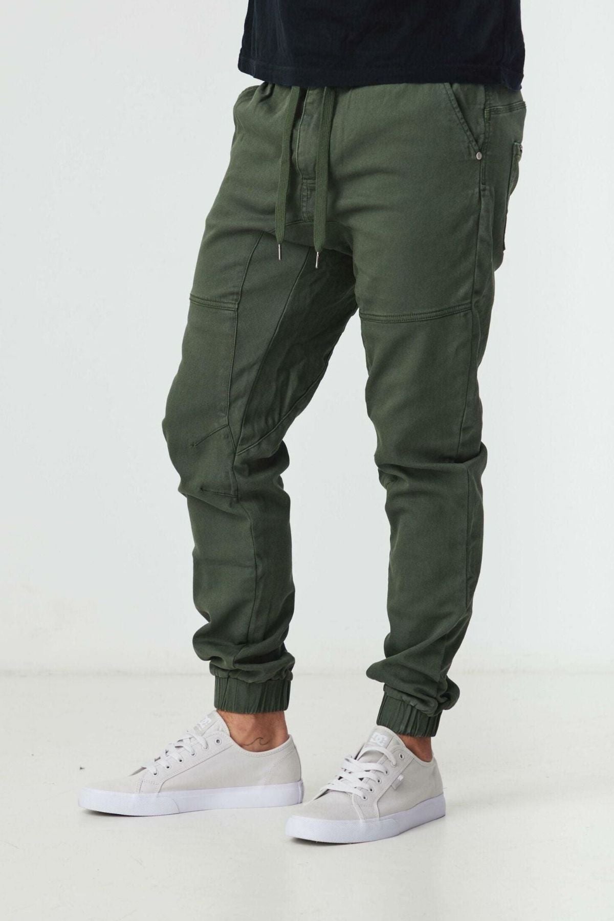 Someone wearing BLKWD James Jogger Pants in olive green, featuring drawstrings and elastic cuffs, pairs them with white sneakers and stands against a plain white background.