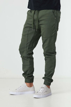 Someone wearing BLKWD James Jogger Pants in olive green, featuring drawstrings and elastic cuffs, pairs them with white sneakers and stands against a plain white background.