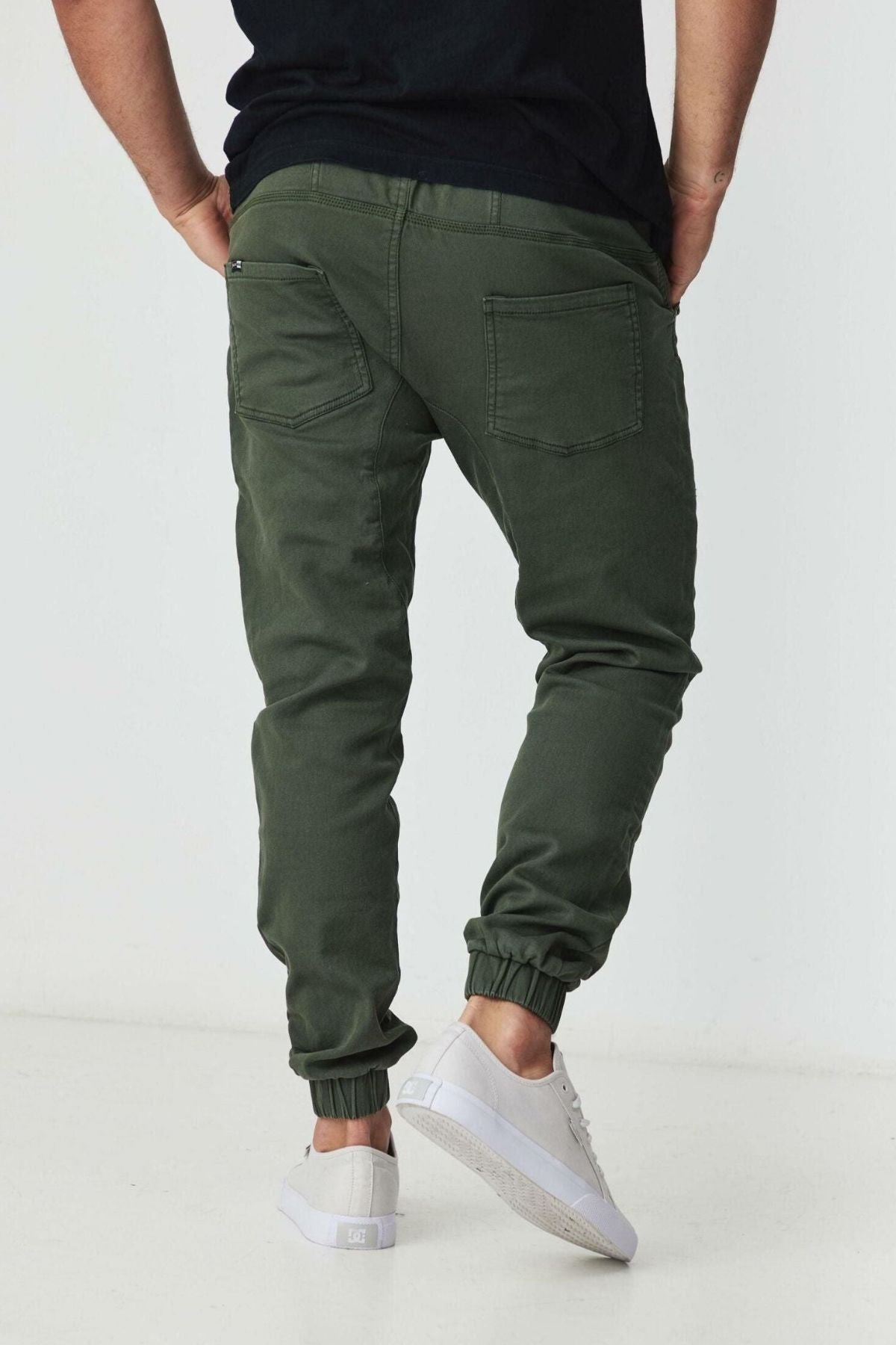 A person wearing BLKWD James Jogger Pants in dark green cotton stretch, a black t-shirt, and white sneakers stands with their back to the camera against a plain white background.