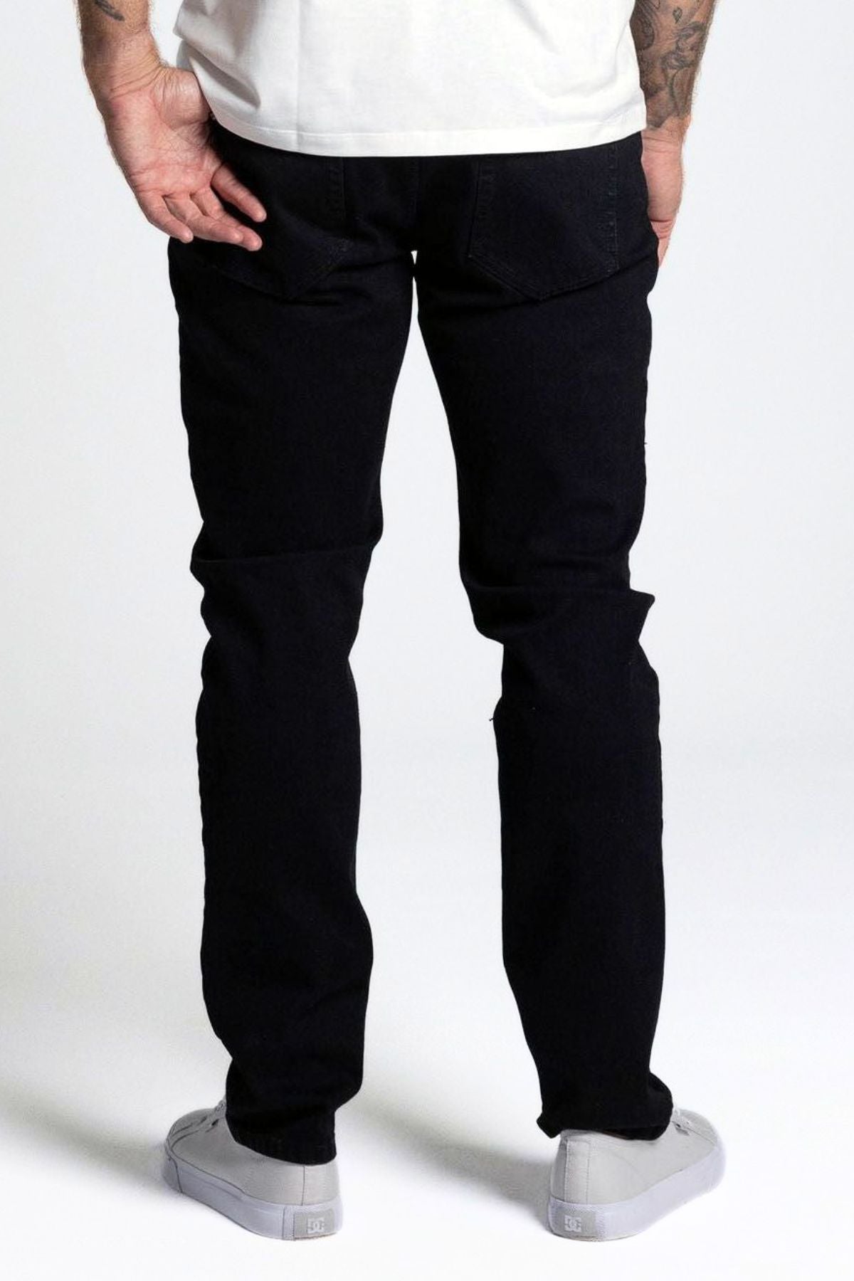 Someone in BLKWD Vassnik Denim Jean Slim Fit, a white shirt, and gray sneakers stands with their back to the camera against a plain light background.