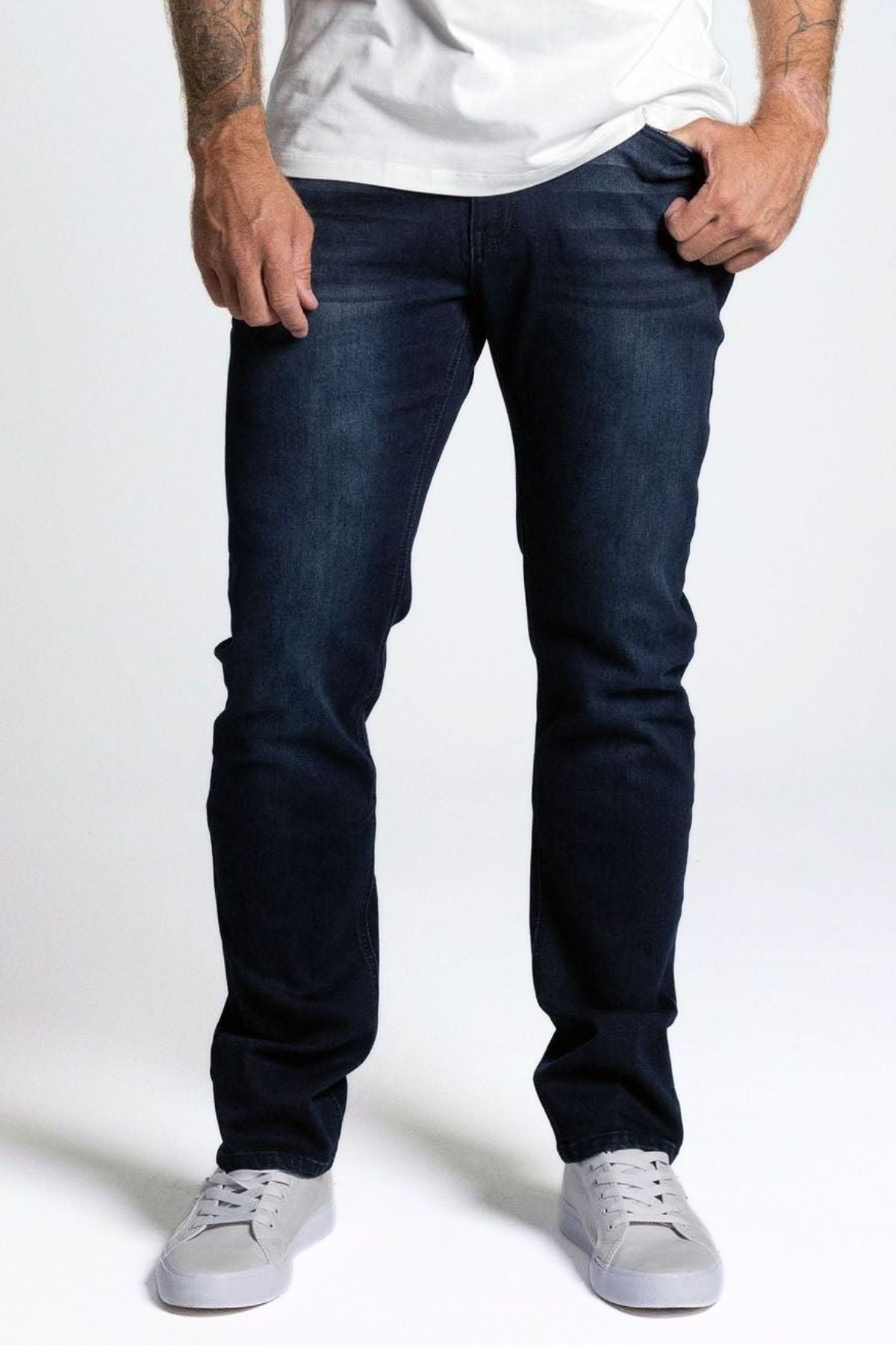 A person in a white T-shirt, BLKWD West Denim Jean Slim with 5-pocket design, and white sneakers stands with hands near pockets, shown from shoulders down against a plain light background.