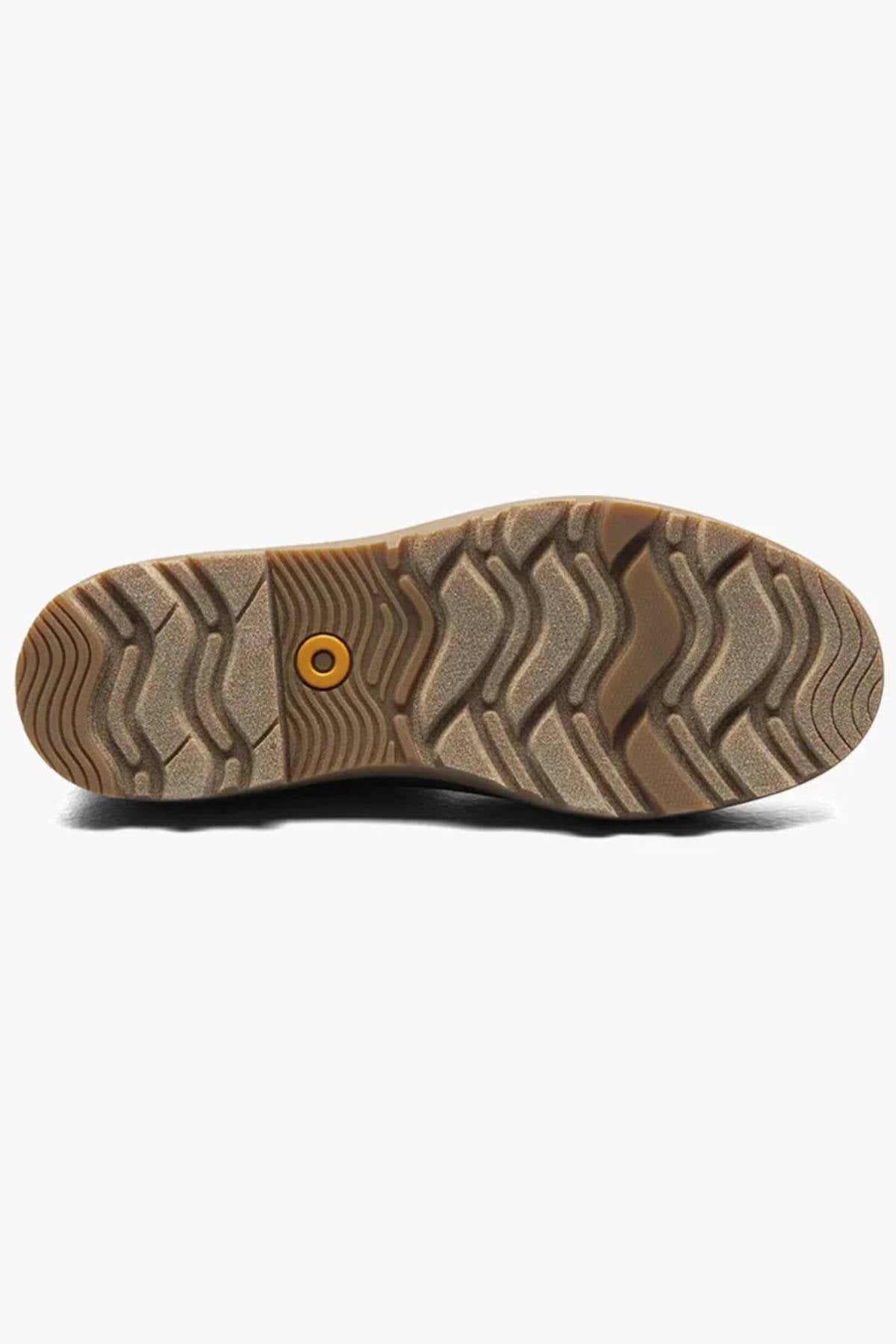 The image shows the sole of the BOGS Holly Chelsea Waterproof Boots, featuring a textured, wavy tread and a circular yellow detail on a light brown rubber surface with BLOOM Algae Footbeds for enhanced comfort.