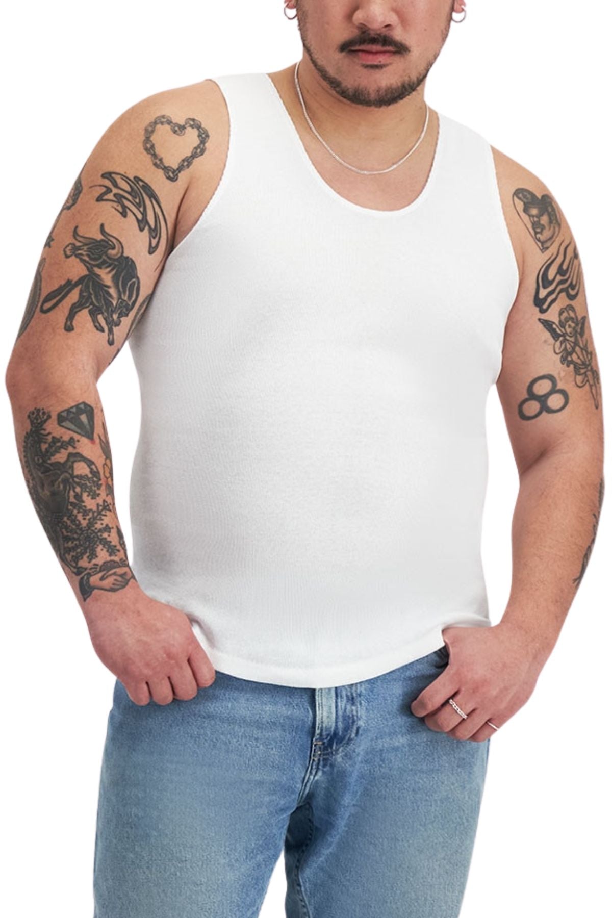 A person models the Bonds Mens Chesty Singlet in white, paired with blue jeans. Tattoos cover their arms and a necklace adorns their neck as they stand against a plain white background.