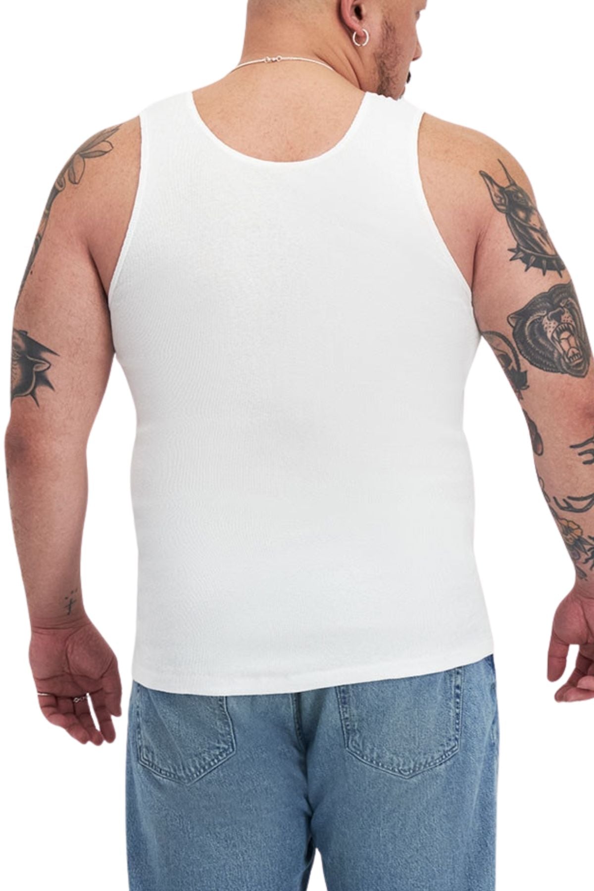 A person models the Bonds Mens Chesty Singlet in white, paired with blue jeans. Tattoos cover their arms and a necklace adorns their neck as they stand against a plain white background.