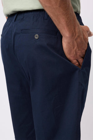 A close-up of a person wearing Breakaway Cody Shorts in navy blue with an elastic waistband, one hand in the side pocket, and a buttoned back pocket. The shorts are ideal for everyday wear and paired with a light green shirt.