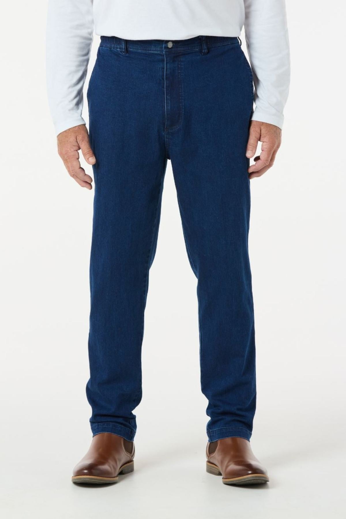 A person wearing a white long-sleeve shirt, the Breakaway Intense Denim Jean with an elastic waist in dark blue, and brown leather shoes is standing. The image shows the person from the neck down on a plain background.