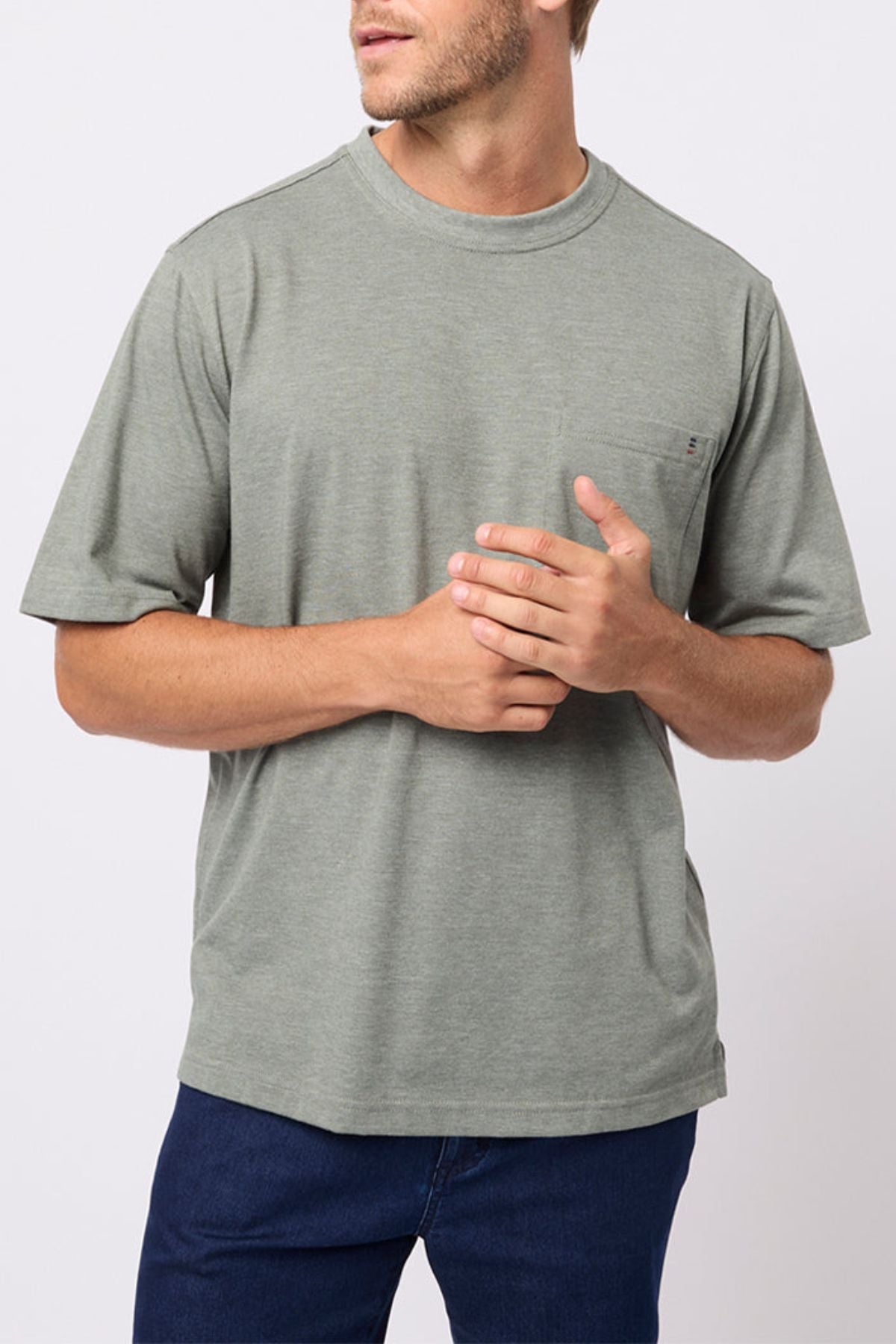 A man in a light gray-green Breakaway Kinnersly Crew, featuring lightweight fabric, a relaxed fit, and chest pocket, stands against a plain background with his hands loosely clasped, looking to the side.