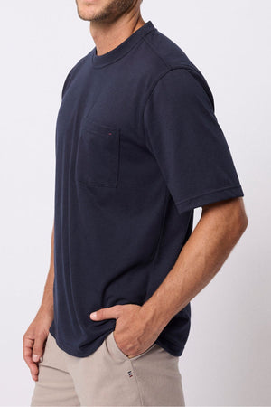 A man in a relaxed fit navy Breakaway Kinnersly Crew t-shirt with a chest pocket and light beige pants stands against a plain light background, one hand in his pocket.