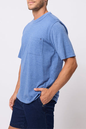 A man stands against a plain white background wearing a relaxed fit Breakaway Kinnersly Crew light blue short-sleeve tee with a chest pocket and dark blue shorts.