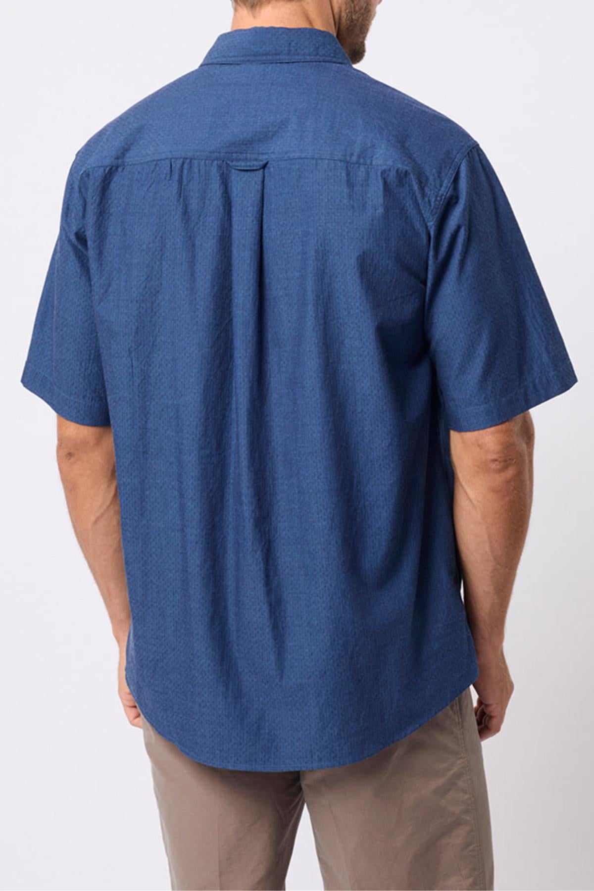 A man is seen from the back wearing the Breakaway Olivero Shirt, a blue short-sleeved 100% cotton men's shirt, paired with light brown pants against a plain light background.