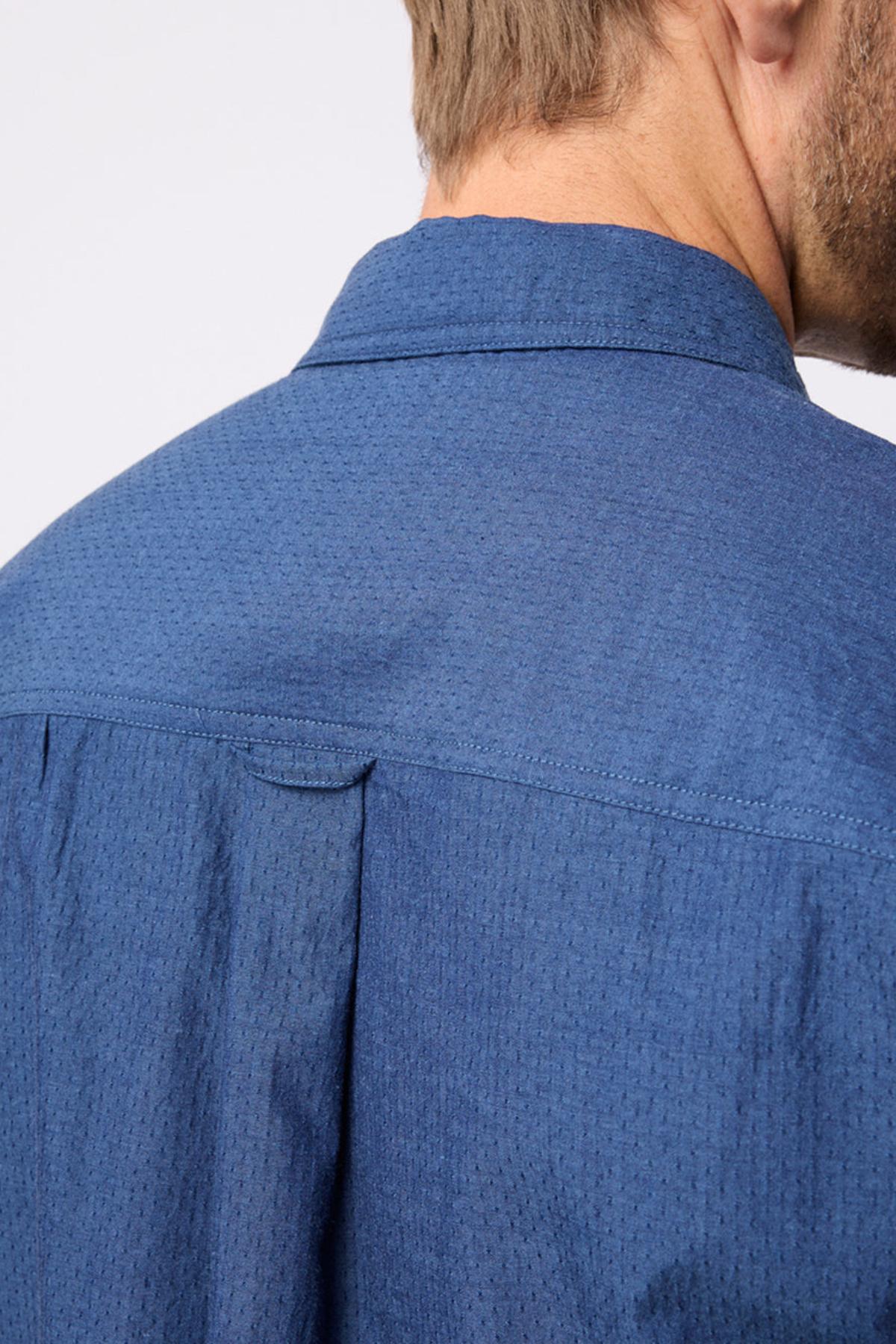 Close-up of a person’s back and shoulder wearing the Breakaway Olivero Shirt. The head is turned away, showing short light brown hair and part of the 100% cotton, textured blue men’s shirt collar.