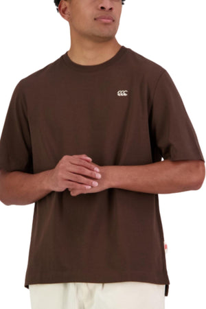 A person wears a brown, oversized Canterbury CCC Collection T-Shirt made from 100% cotton with a small white chest logo, paired with light-colored pants, standing with hands clasped in front.
