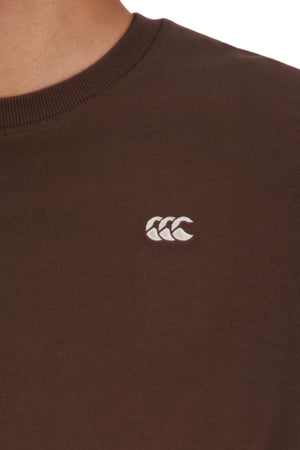 Close-up of a person wearing the oversized Canterbury CCC Collection T-Shirt in dark brown, made from 100% cotton, and featuring a small white embroidered logo with three overlapping curved shapes on the chest.