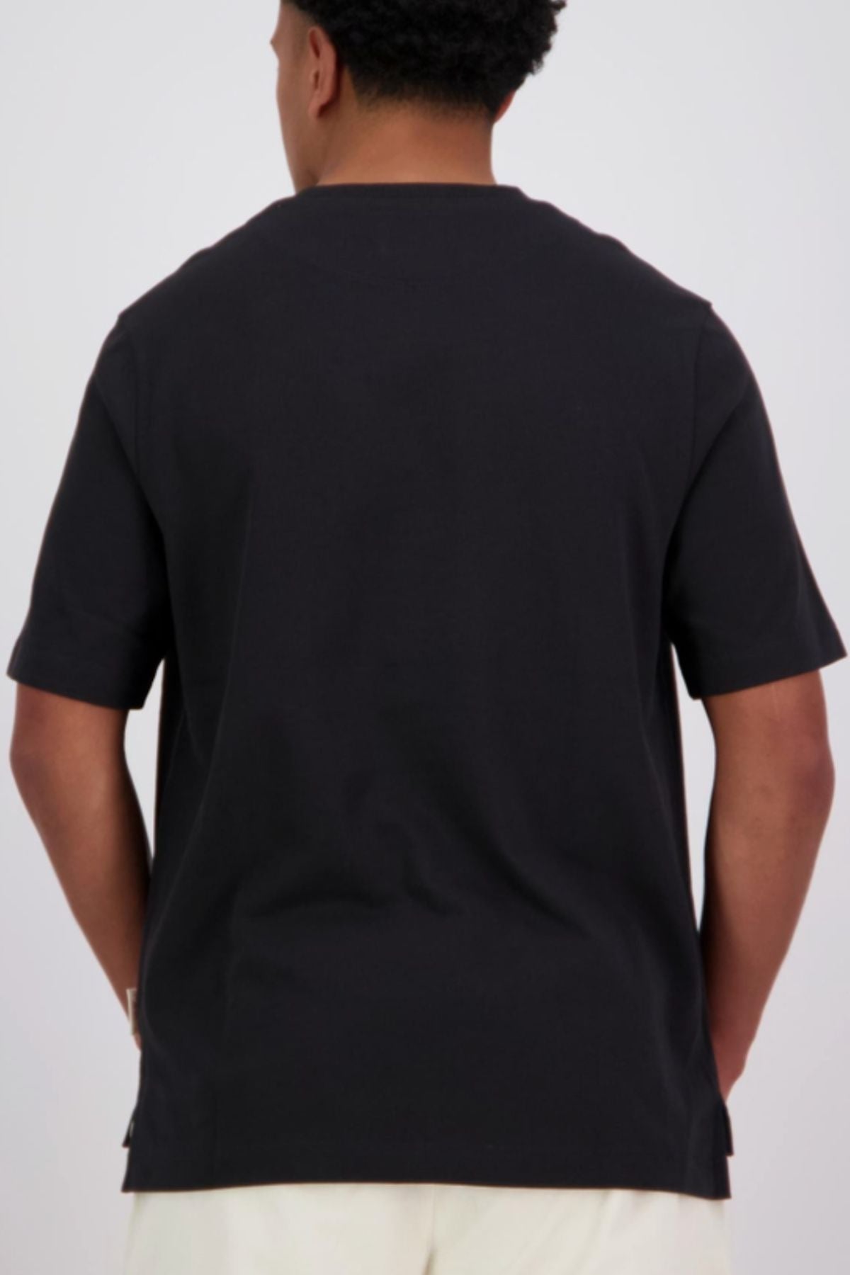 A person faces away from the camera, wearing a black oversized Canterbury CCC Collection T-Shirt and light-colored pants against a plain background.