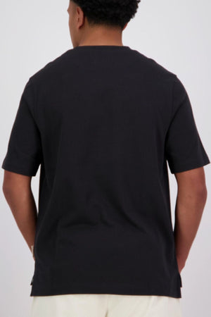 A person faces away from the camera, wearing a black oversized Canterbury CCC Collection T-Shirt and light-colored pants against a plain background.