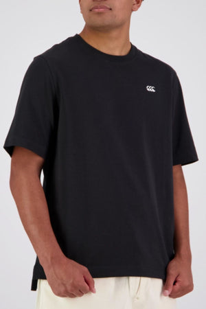 A person wearing a loose-fitting Canterbury CCC Collection T-Shirt in black with a small white chest logo and light-colored pants, standing against a plain background.