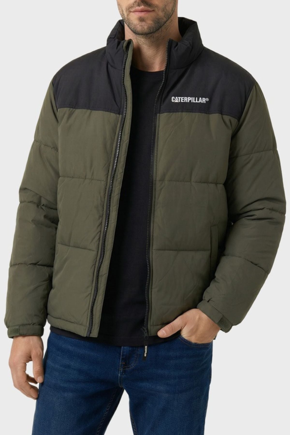 A man stands with one hand in his pocket against a plain light background, dressed in a green and black CAT Heavy Weight Insulated Puffer Jacket with recycled insulation, complemented by a black shirt and blue jeans.