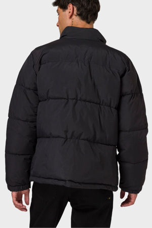 A person is shown from behind, looking slightly to the side against a plain light gray background, wearing the CAT Heavy Weight Insulated Puffer Jacket and black pants.