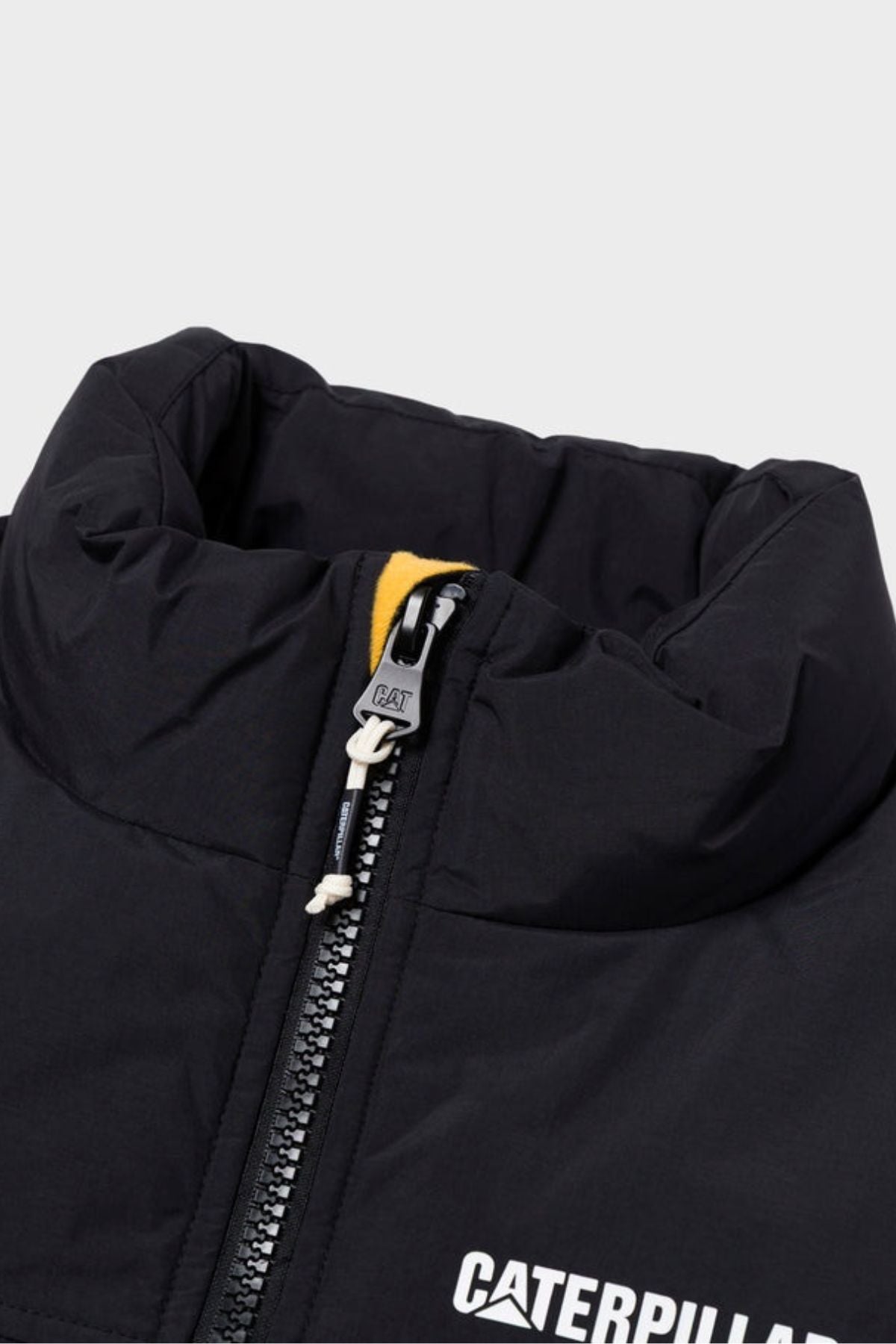 Close-up of the CAT Heavy Weight Insulated Puffer Jacket in black, featuring a visible zipper, yellow fabric detail underneath, and partially visible white "CATERPILLAR" lettering on the chest.