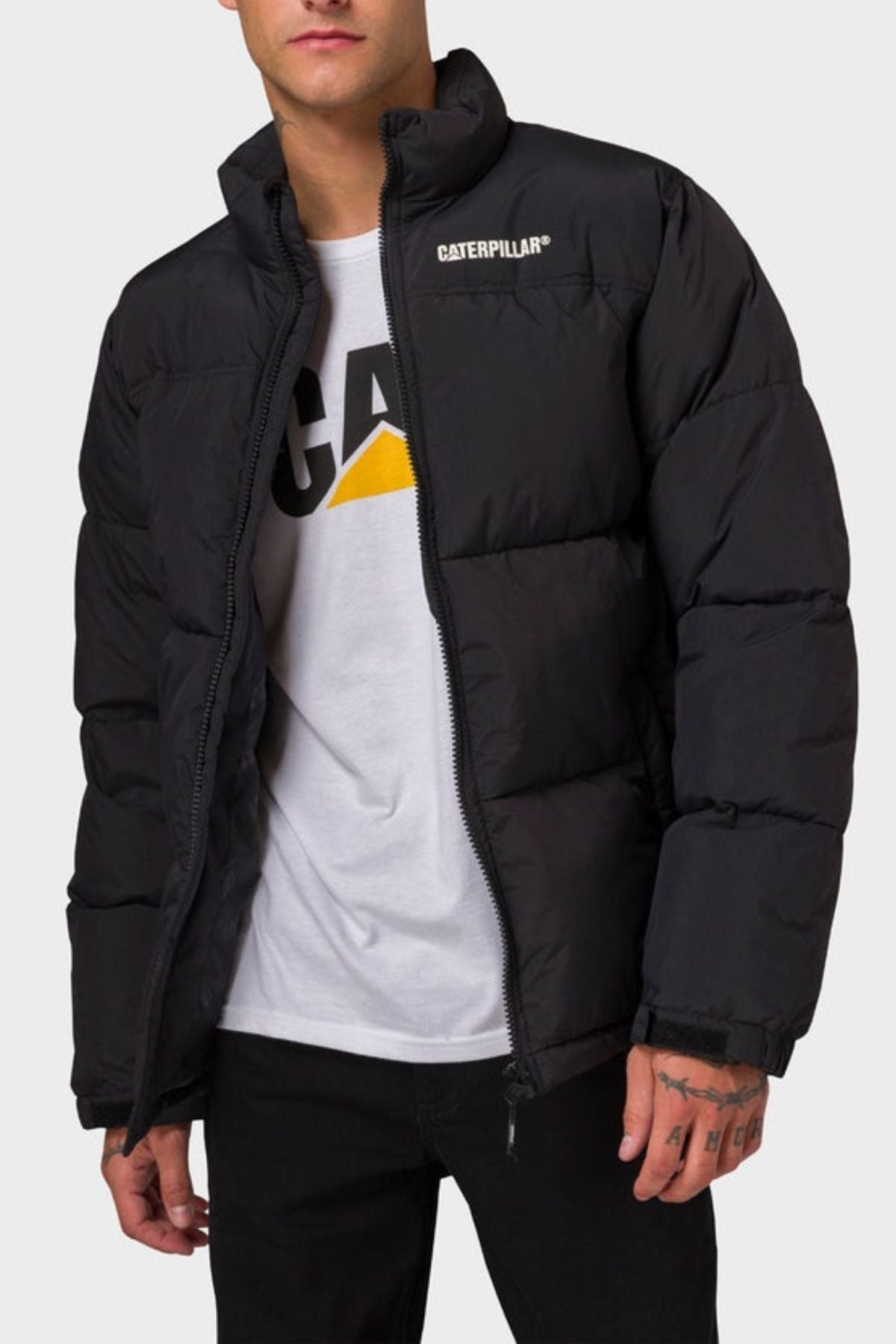 A person wearing the CAT Heavy Weight Insulated Puffer Jacket over a white T-shirt with a yellow and black graphic, paired with black pants, stands against a plain light background.