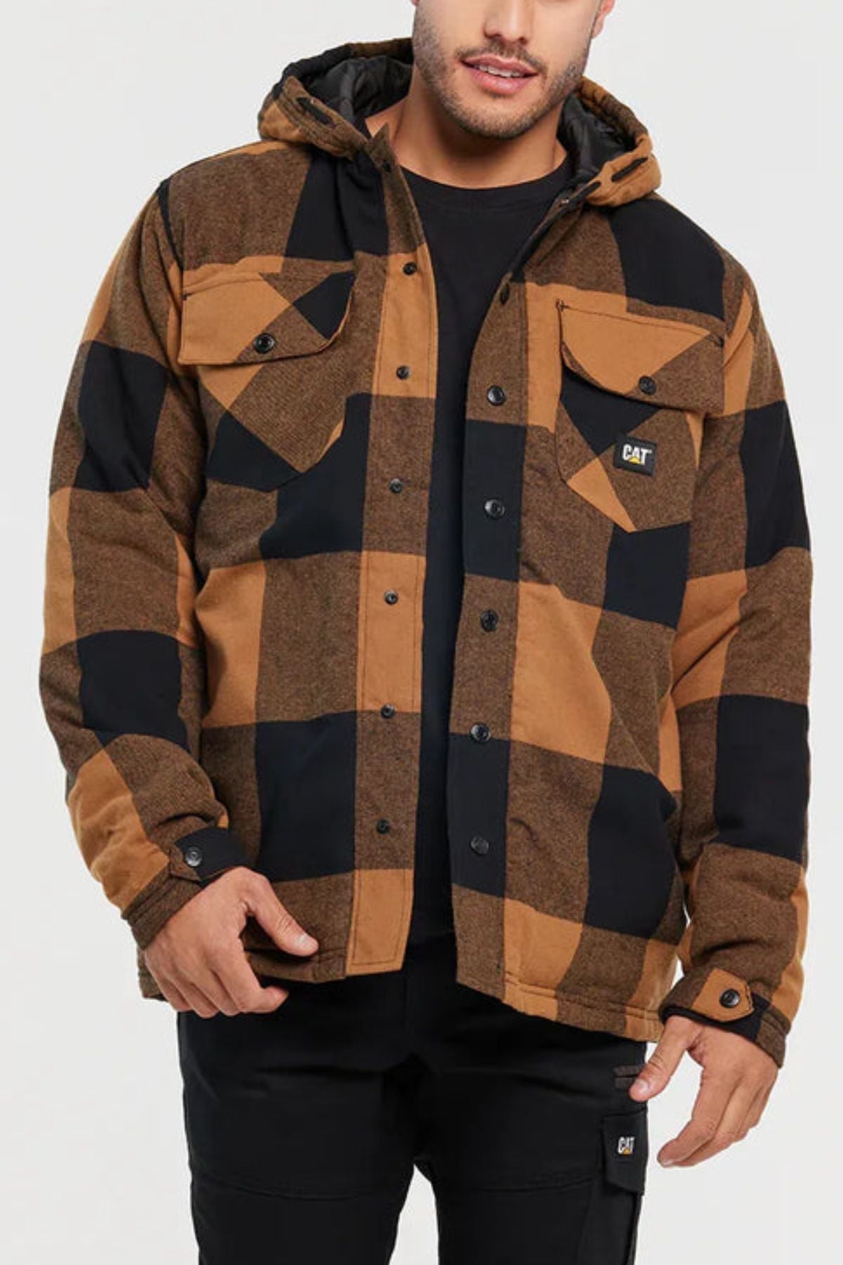 Wearing a brown and black plaid CAT Hooded Flannel Shirt Jacket with black buttons, chest pockets, and black pants, a person stands against a plain light background. The CAT Hooded Flannel Shirt Jacket combines comfort and style.