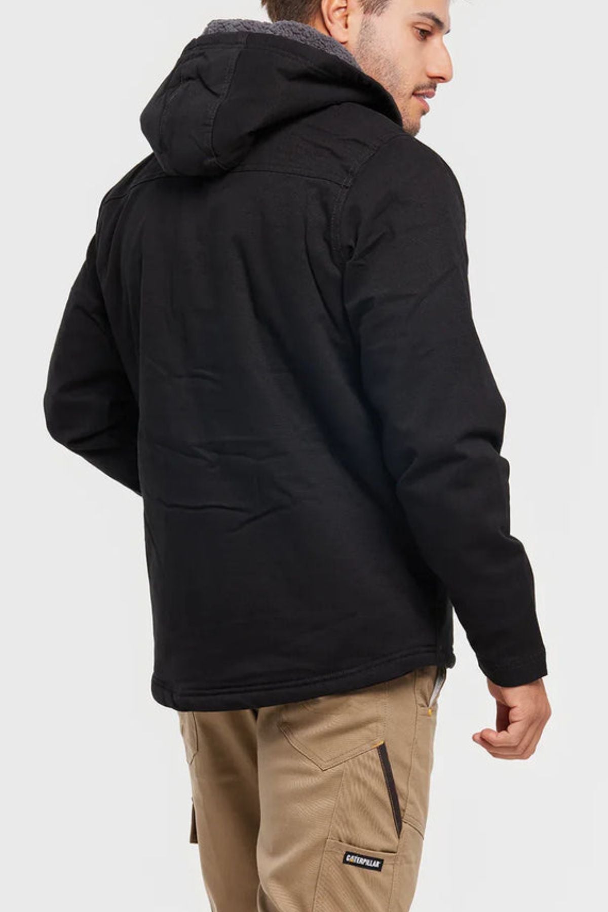 A man in a CAT Hooded Sherpa Lined Jacket and light brown pants stands facing away, turning slightly to the side. The jacket has a hood, and the pants feature zippered pockets with a "Caterpillar" logo.
