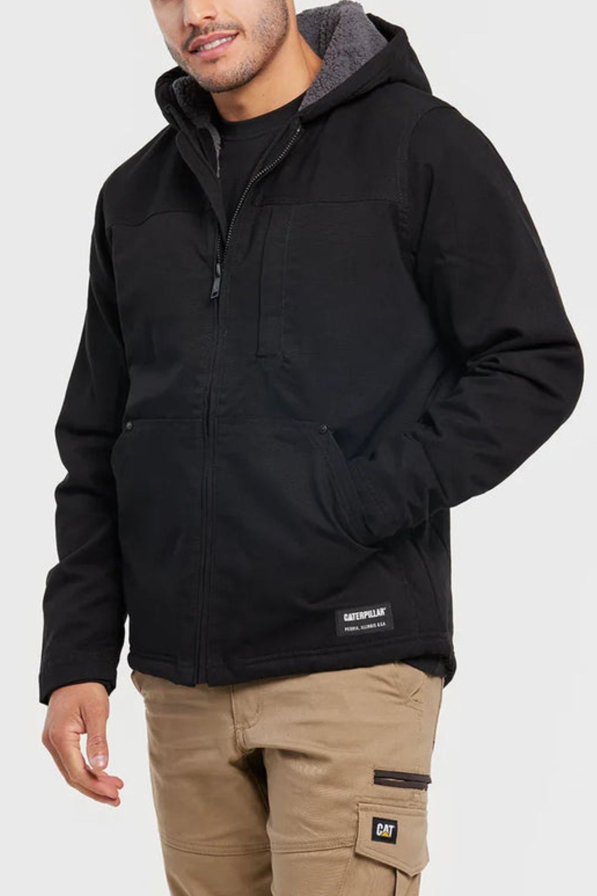 A person wearing a black CAT Hooded Sherpa Lined Jacket with a gray lining and tan work pants with pockets and a “CAT” logo patch, standing against a plain light background.