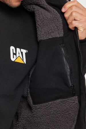 Close-up of a person wearing the CAT Hooded Sherpa Lined Jacket in black with gray fleece, holding it open to reveal an inner zippered pocket. The shirt underneath features the white and yellow CAT logo.