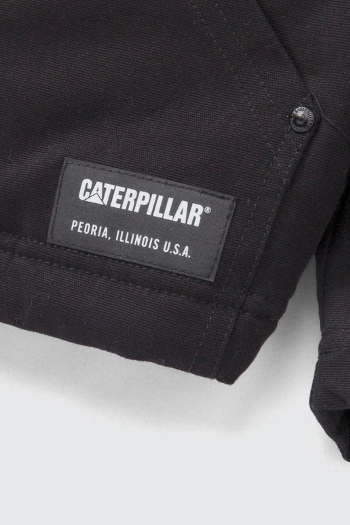 Close-up of a black fabric label stitched onto the CAT Hooded Sherpa Lined Jacket, featuring the "CATERPILLAR" logo in white text above "PEORIA, ILLINOIS U.S.A.