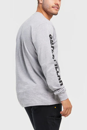 A man in a comfortable CAT Trademark Banner Long Sleeve Tee with black sleeve text and black pants stands sideways, looking over his shoulder against a plain white background.
