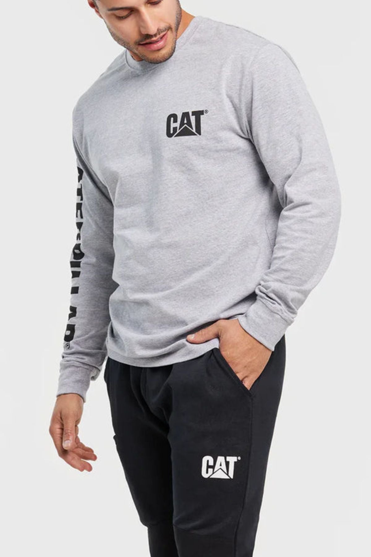 A man in a comfortable CAT Trademark Banner Long Sleeve Tee with "CATERPILLAR" on the sleeve and black CAT sweatpants stands with one hand in his pocket, looking downward.