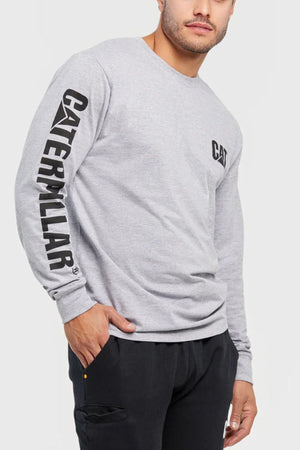 A man wears a light gray CAT Trademark Banner Long Sleeve Tee with "CATERPILLAR" on the right sleeve and "CAT" on the chest, paired with black pants.
