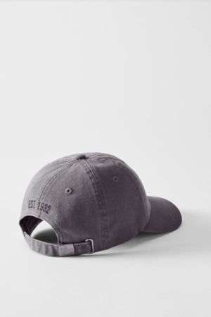 A gray RM Williams Mini Longhorn Twill Cap is positioned backwards on a plain white surface, displaying its adjustable strap and the small "EST. 1932" embroidery on the back.