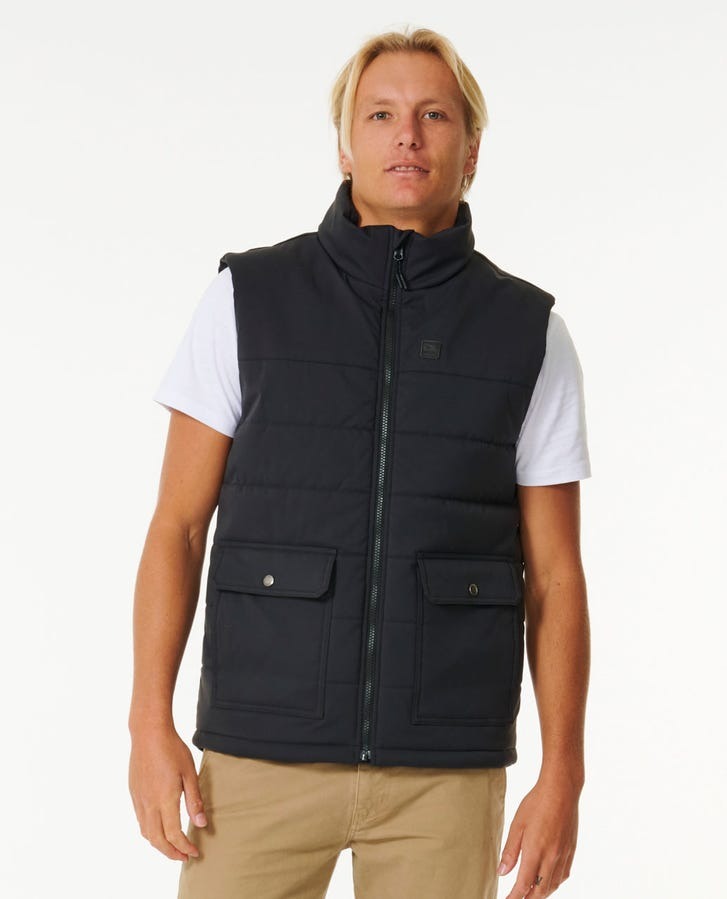A man with blonde hair stands facing the camera, clad in a Rip Curl Ridge Anti Series Vest over a white T-shirt and khaki pants. The durable, water-resistant vest boasts two front pockets secured with button flaps. The background is plain white.