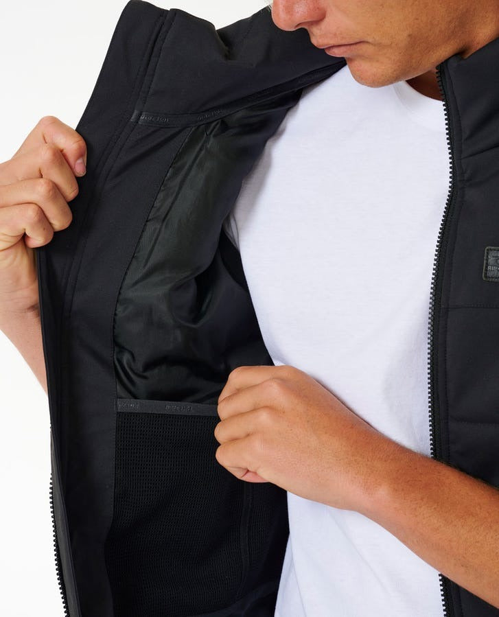 An individual is unzipping the front of a black Rip Curl Ridge Anti Series Vest, revealing the inner pockets. They are dressed in a white t-shirt beneath the vest. The person’s face is partially visible, looking downward at the winter garment against a plain white background.