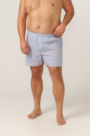 A barefoot man with a tattoo on his left forearm models the Contare Cotton 2 Pack Check Boxer Short in blue and orange plaid, standing against a plain light background with his bare torso and right hand near the waistband.