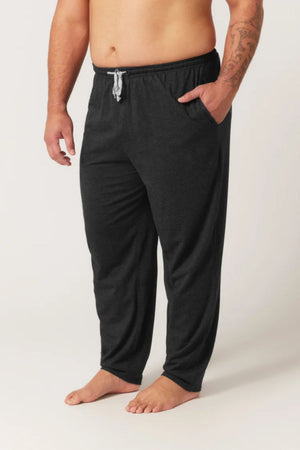 Someone stands barefoot against a plain background, wearing Contare Country Bamboo Cotton Lounge Pants in dark gray with a light gray drawstring, one hand in their pocket.