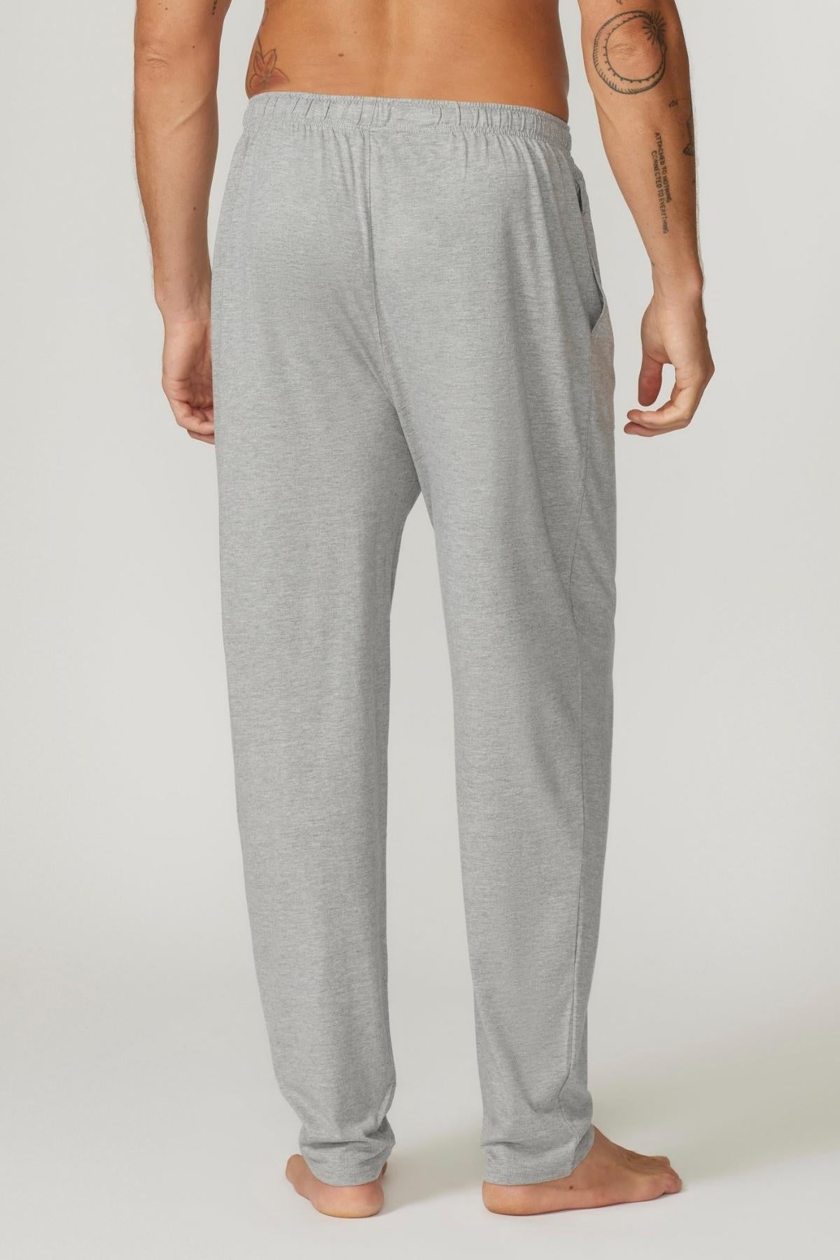 A person standing barefoot, facing away from the camera, wearing Contare Country Bamboo Cotton Lounge Pants in light gray. Their left arm, adorned with tattoos on the arm and shoulder, hangs by their side. The lounge pants are loose-fitting and appear to have pockets on both sides.