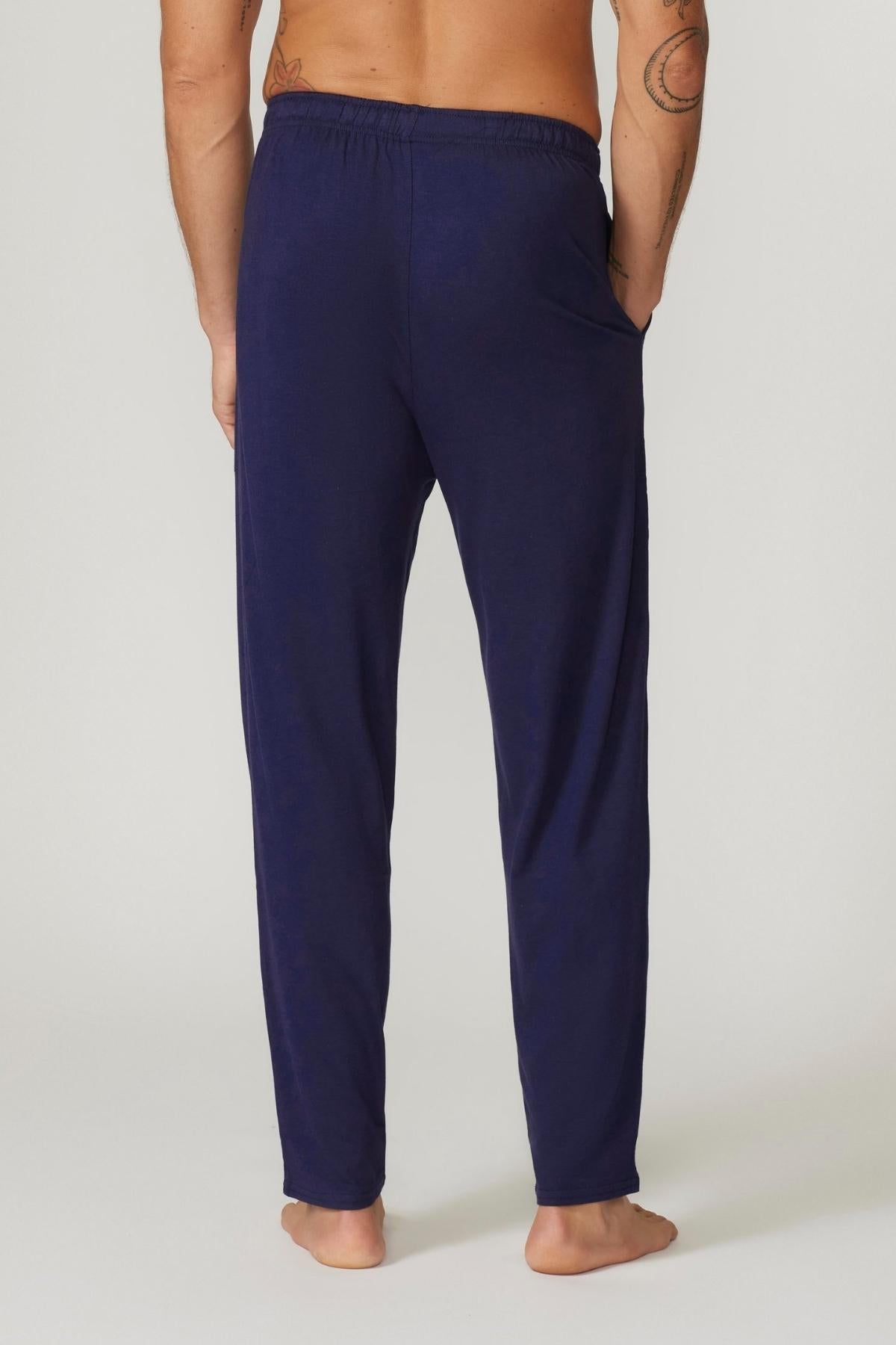 A person stands barefoot on a light surface, shown from the back, wearing dark blue Contare Country Bamboo Cotton Lounge Pants with hands in side pockets. The breathable and eco-friendly fabric complements shirtless comfort and visible arm tattoos.