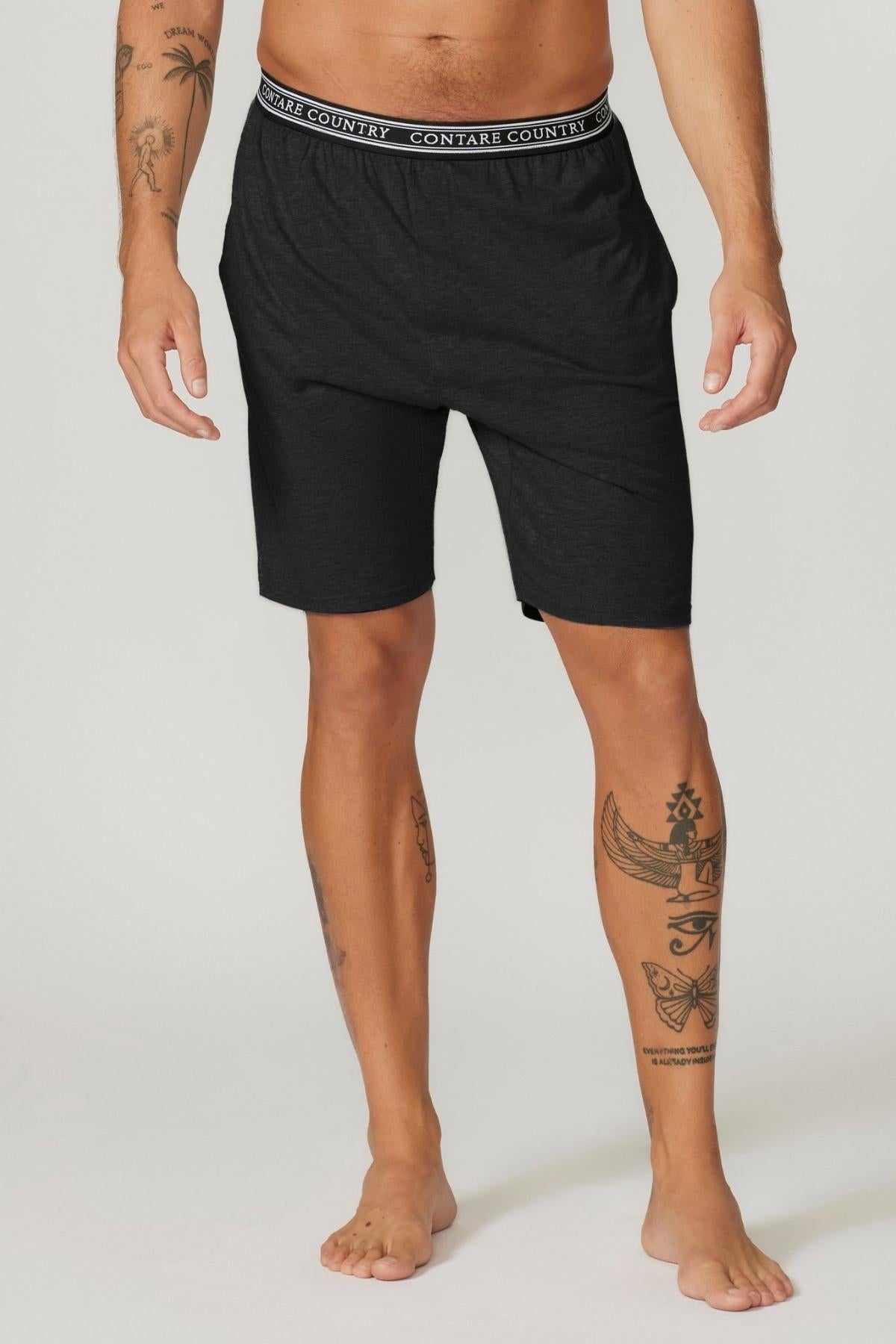 A person is standing barefoot, wearing black Contare Country Bamboo Cotton Sleep Short with "Contare Country" written on the waistband. They have various tattoos on their arms and legs. The background is plain and light-colored. The person's head is not visible in the image.