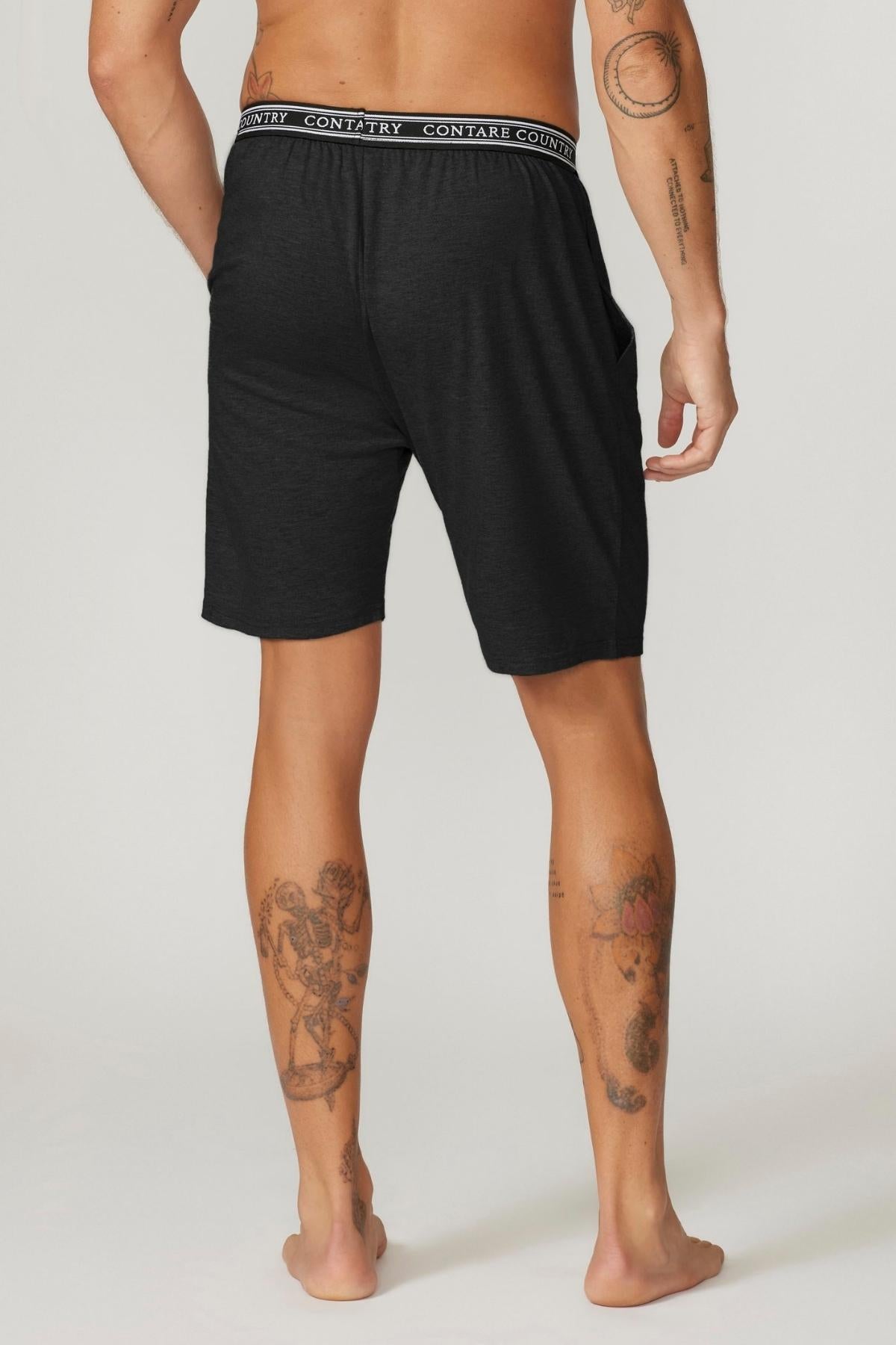 A person with visible tattoos on their arms and legs is standing barefoot, facing away. They are wearing Contare Country Bamboo Cotton Sleep Shorts in black, which feature an elastic waistband with a repeating "CONTRARY" text. Their hands are resting casually at their sides.
