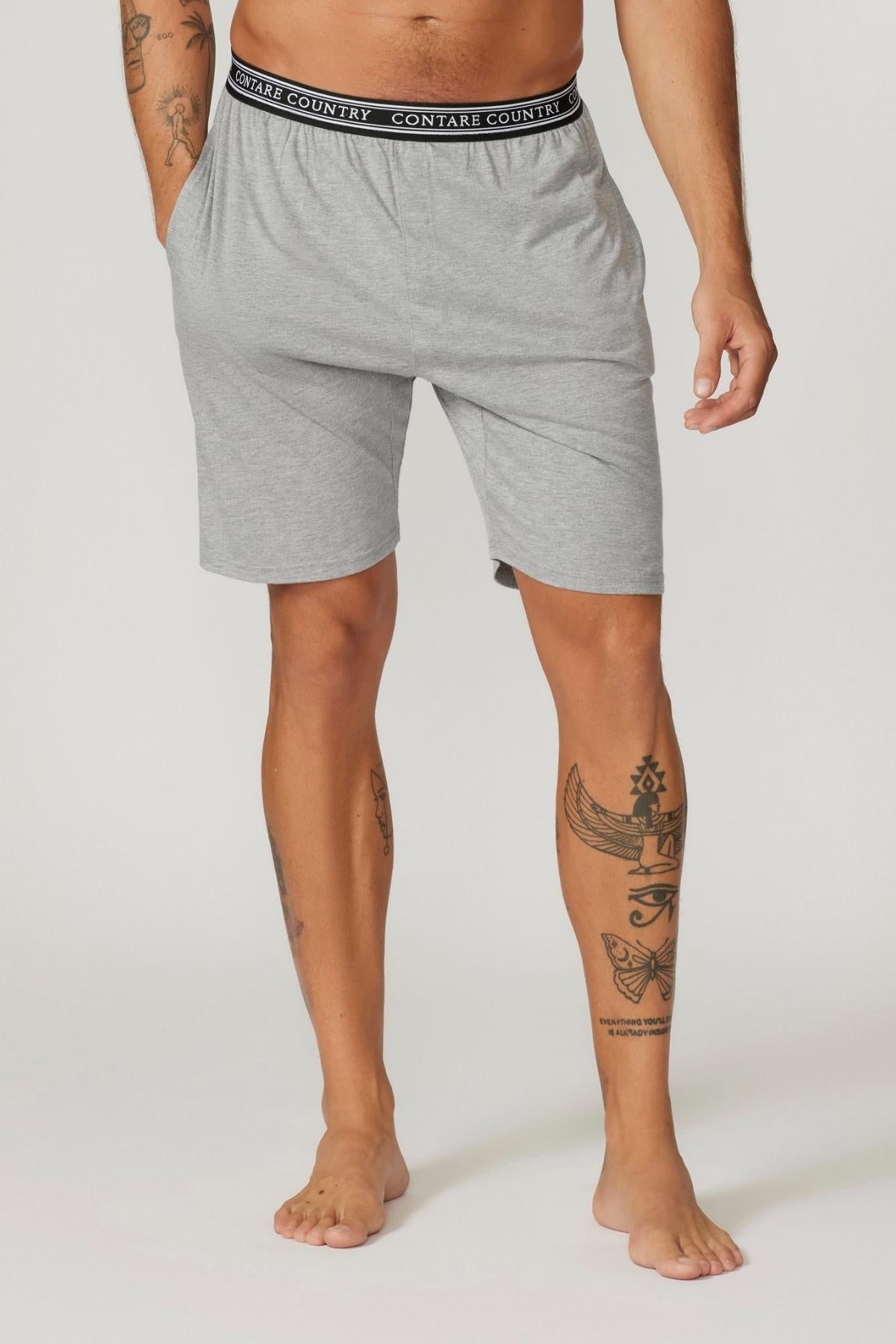 A person with visible tattoos on their arms and legs is shown from the neck down, wearing Contare Country Bamboo Cotton Sleep Shorts. The shorts are light gray with a black waistband featuring white text that reads "CONTARE COUNTRY." Their hands are in the pockets, and they are barefoot.