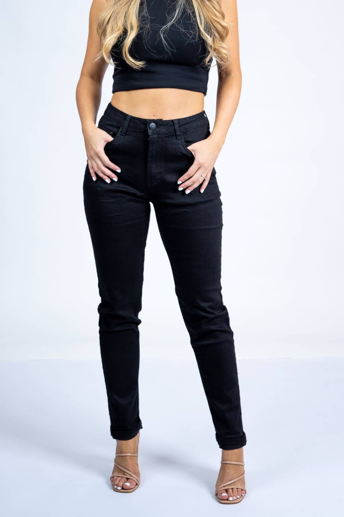 A woman stands against a plain white background wearing a black sleeveless crop top, Country Denim Relaxed Mum Jean high-waisted black jeans, and nude strappy heels. Her hands are in her pockets, with her head out of frame.
