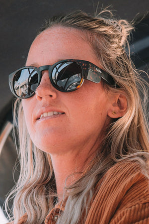 A woman with long blonde hair wears SafeStyle Cruisers Matte Brown Tort sunglasses with brown polarised lenses, looking to the side and smiling. Her sunglasses reflect a car and street. She pairs them with a brown jacket.